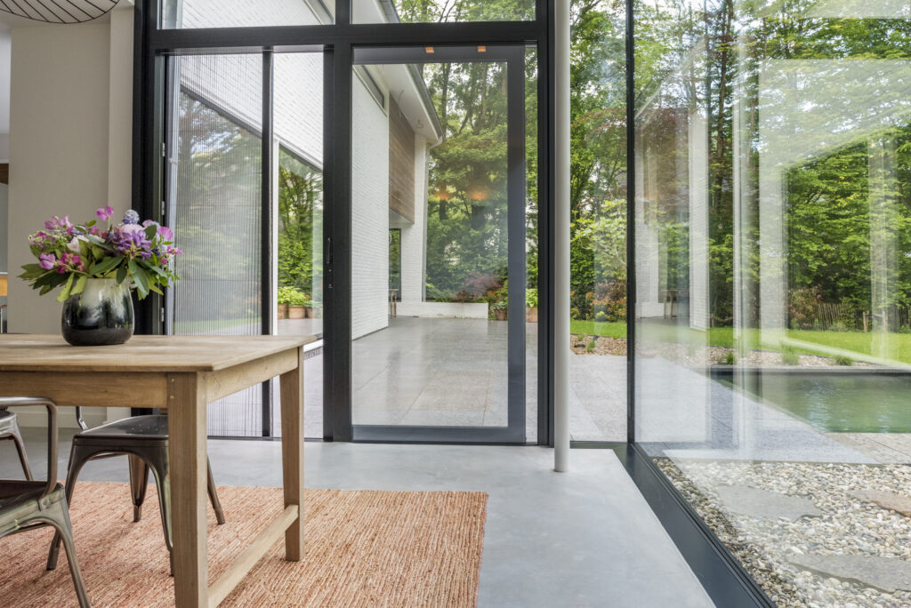 Licht en modern interieur met grote aluminium schuiframen die uitkijken op een terras en tuin met zwembad, ontworpen voor maximale lichtinval en een naadloze overgang tussen binnen en buiten.