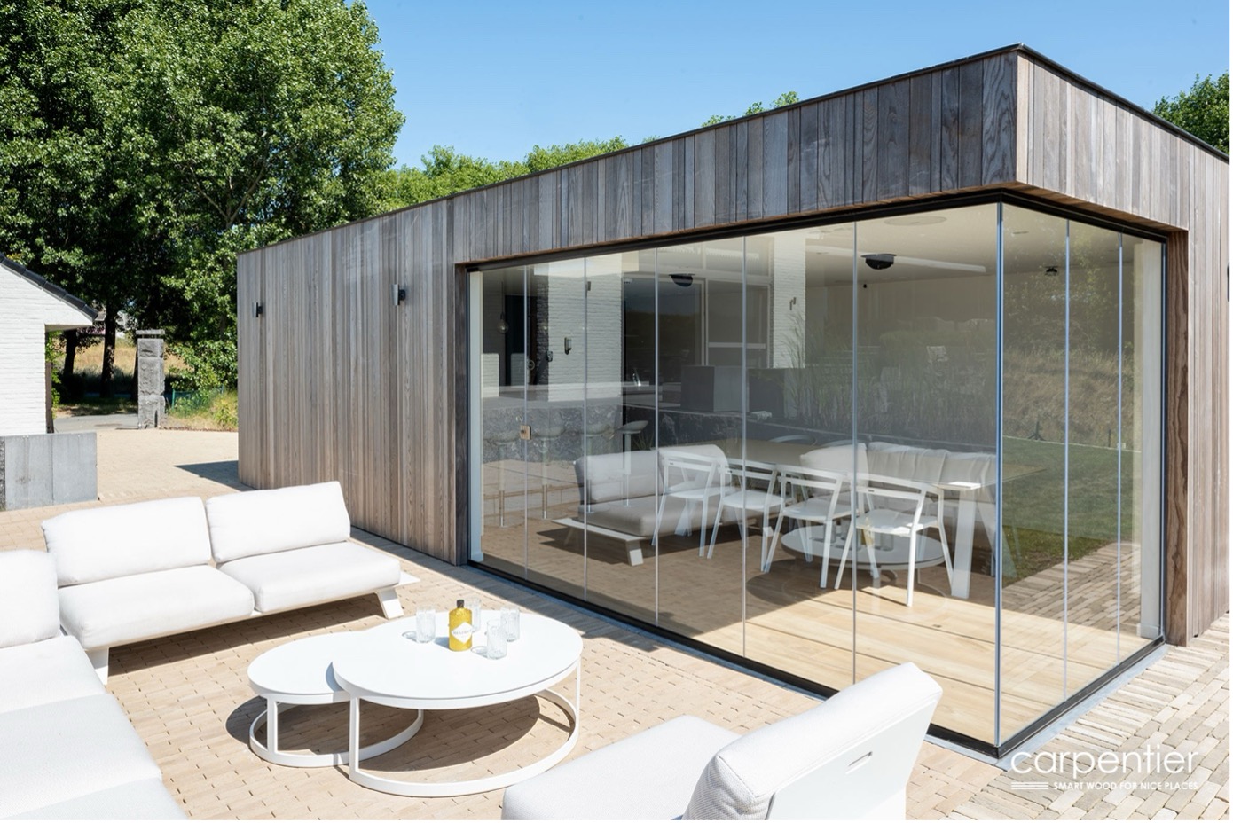 Strak houten bijgebouw met grote glazen schuifwanden, uitkijkend op een terras met wit buitenmeubilair en omringd door groen.