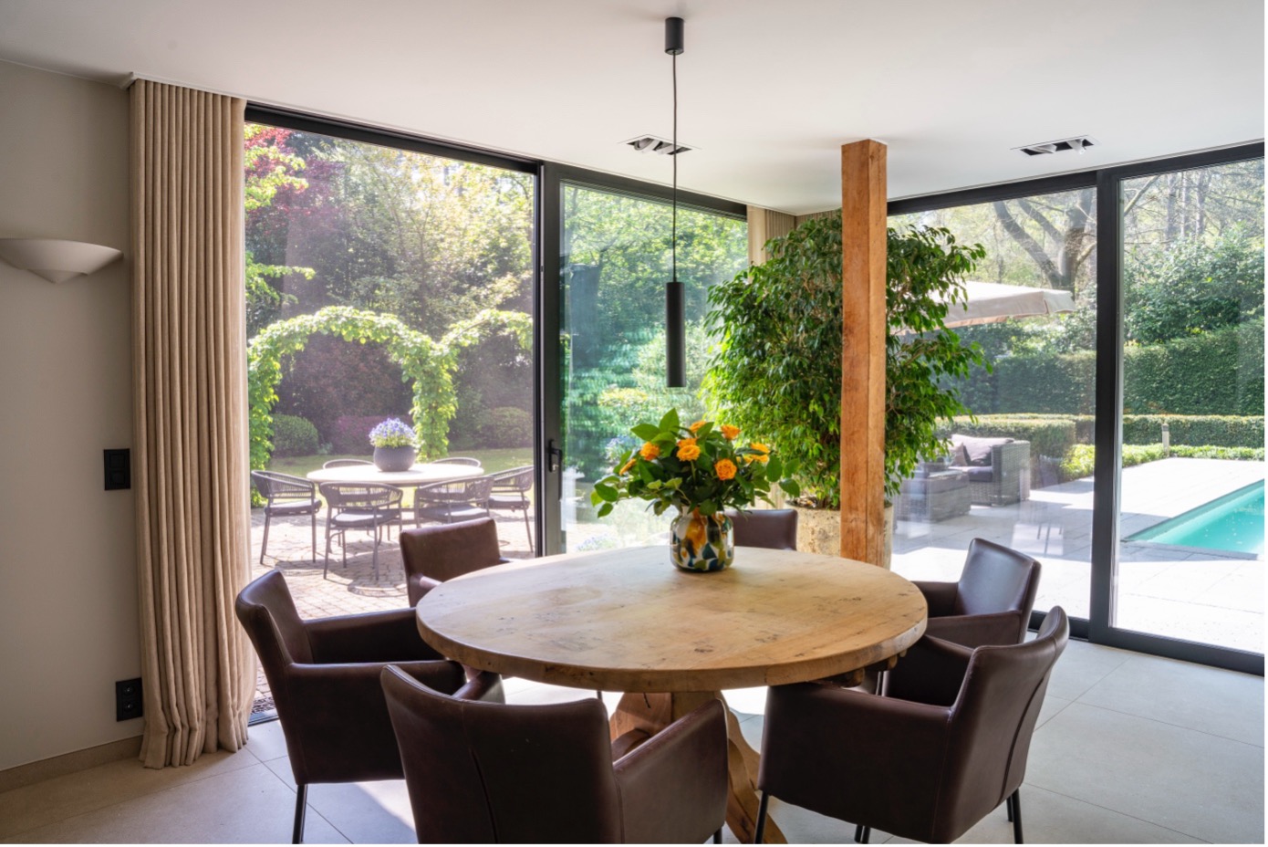 Eetruimte met grote glazen schuiframen die uitkijken op een zonnige tuin met pergola en zwembad, ingericht met een ronde houten tafel en lederen stoelen.
