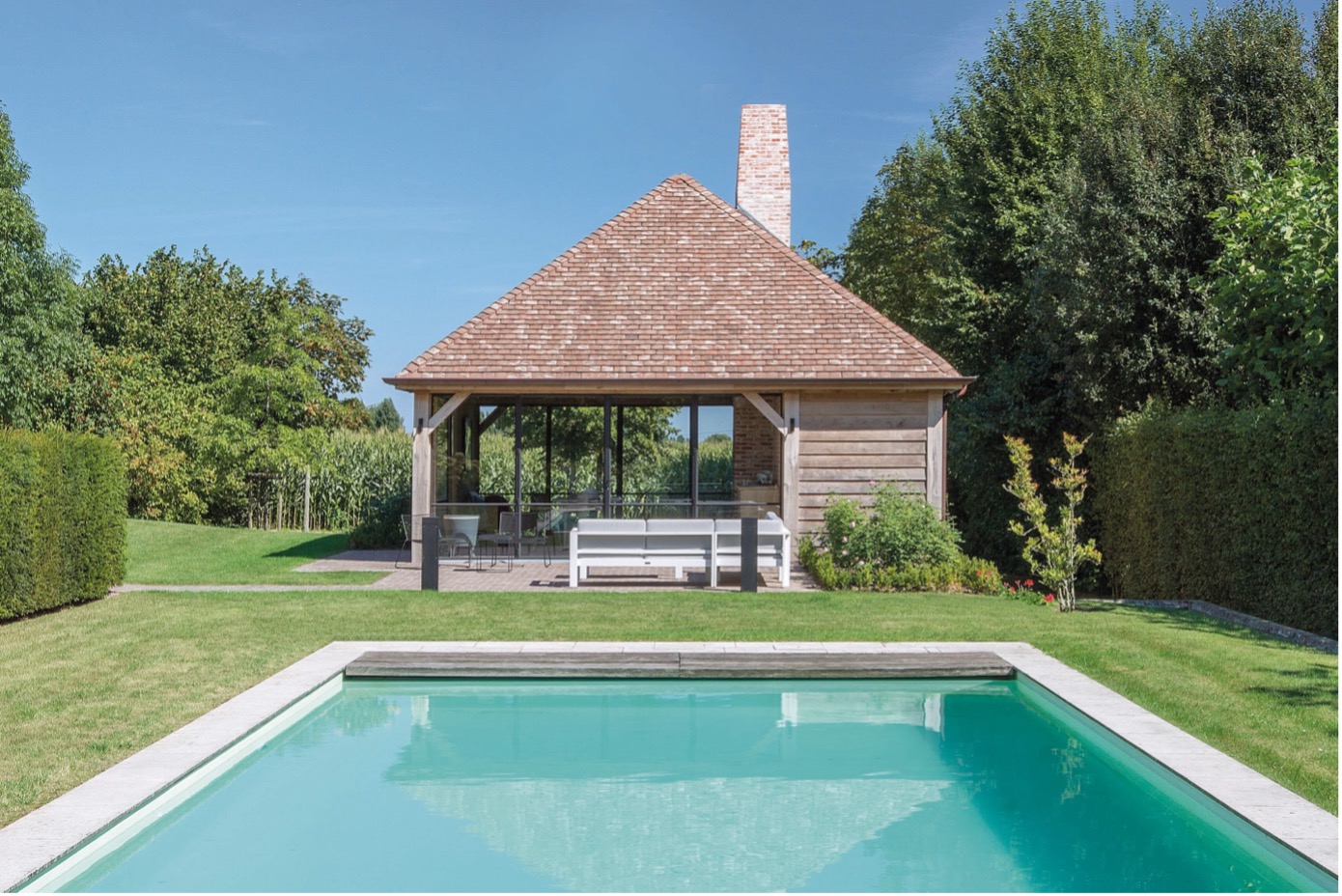 Houten poolhouse met grote glaspartijen en terrasmeubilair, gelegen in een groene tuin met een strak aangelegd zwembad op de voorgrond.