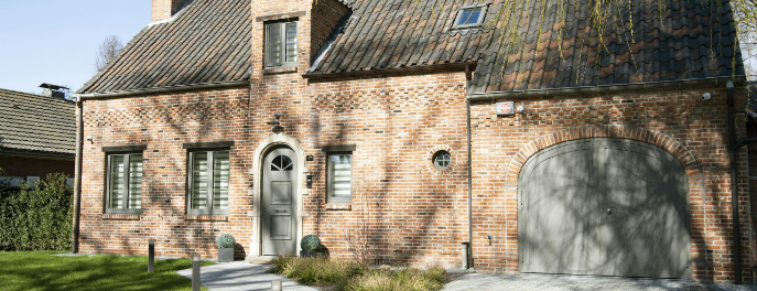 Landelijke woning met aluminium retro voordeur en bijpassende garagepoort in taupe, geïntegreerd in een klassieke rode bakstenen gevel met authentiek dak.