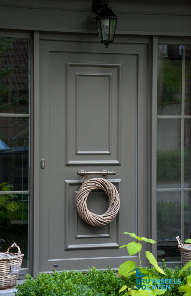 Softline 82 voordeur in klassieke stijl, uitgevoerd in taupe met sierpanelen en decoratieve krans, geplaatst door Bruynseels-Vochten.