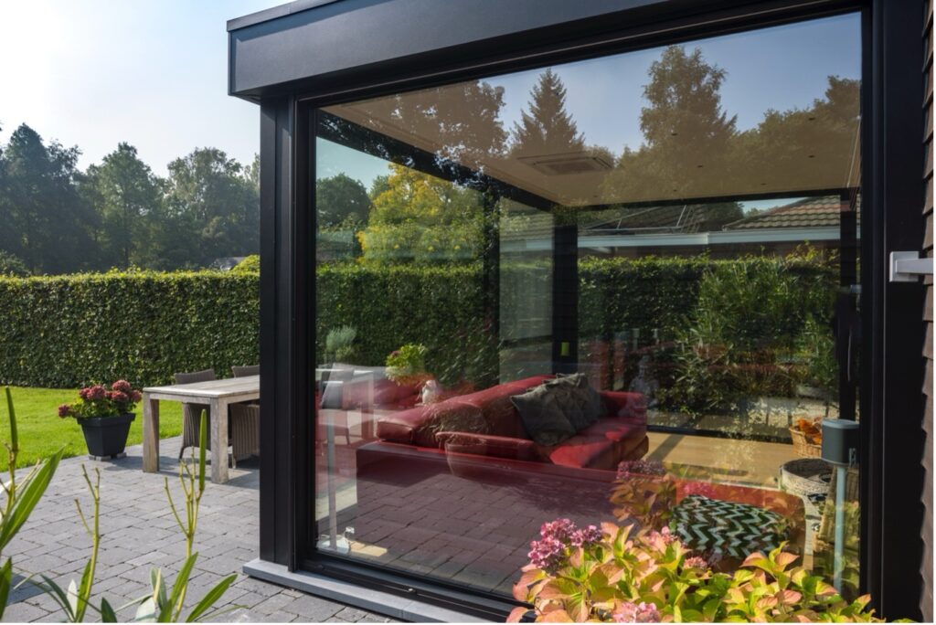 Moderne veranda met grote glaspartijen en zicht op een rood lounge-salon, omringd door een verzorgde tuin met terras en groen.